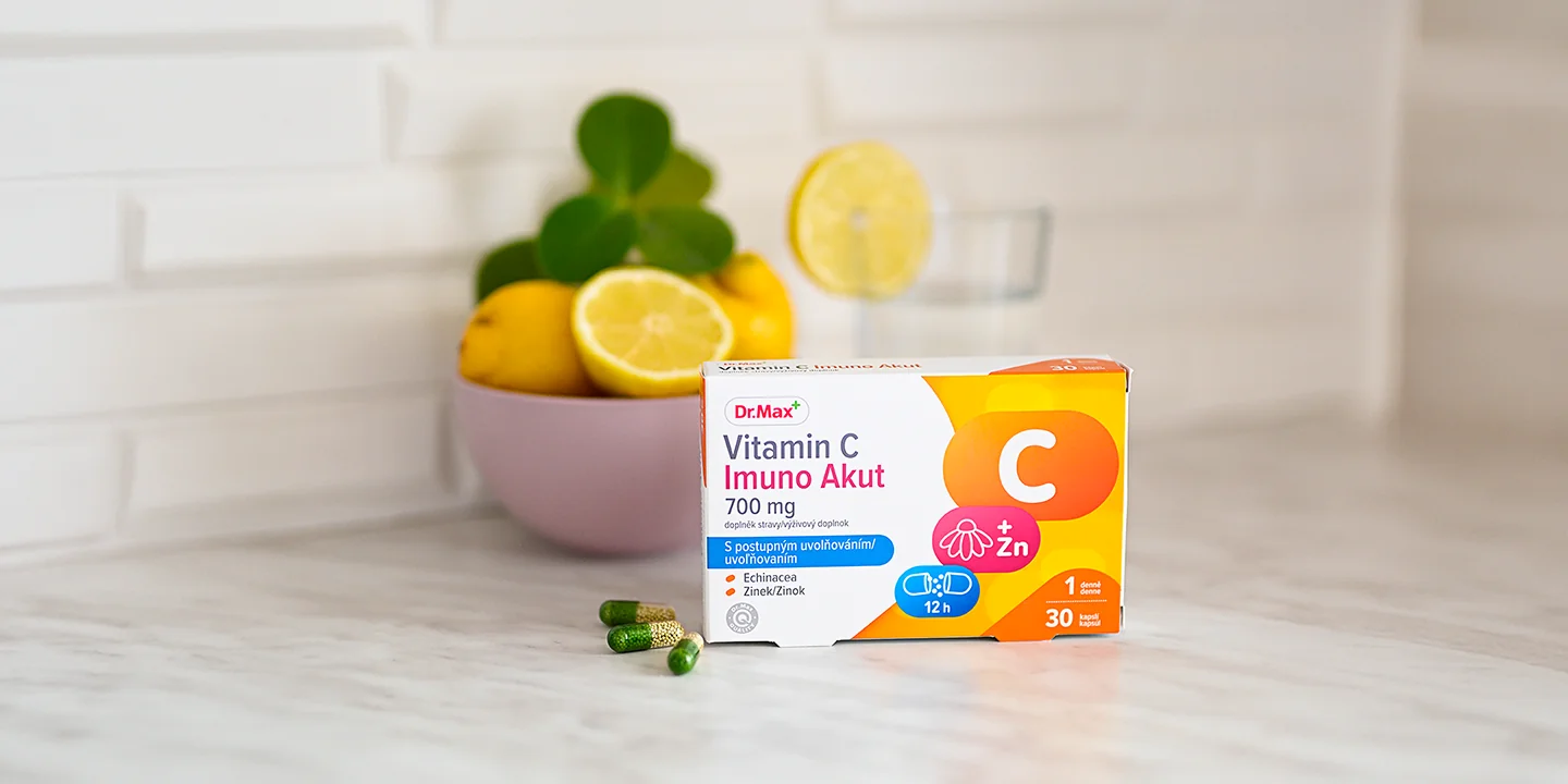 Výživový doplnok Dr. Max Vitamin C Imuno Akut 700 mg v krabičke s voľne položenými kapsulami na kuchynskej linke, v pozadí s miskou plnou čerstvých citrónov a pohárom vody ako ukážka efektívneho dopĺňania vitamínu C pre zdravie a imunitu.